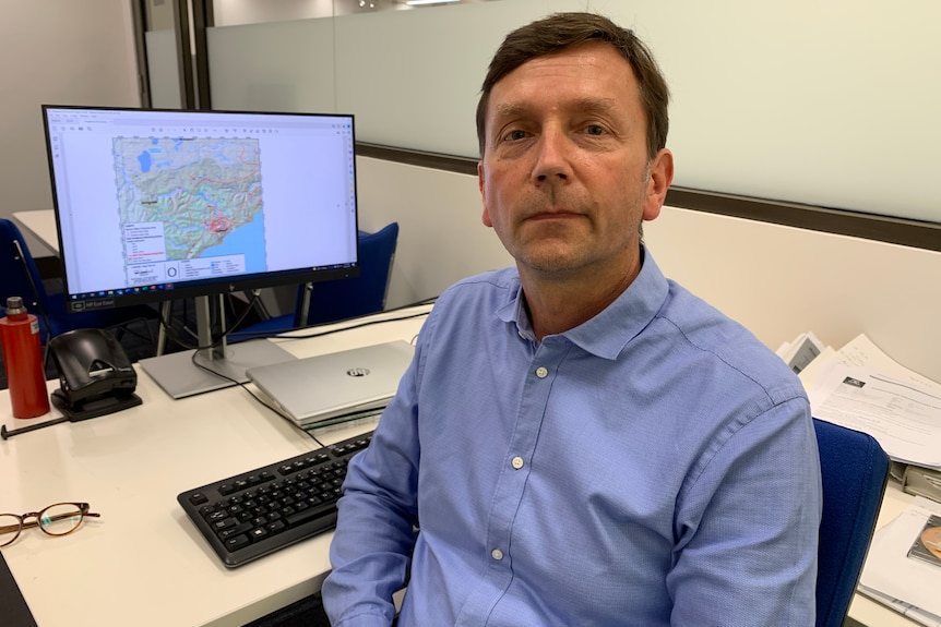 Ralf Haese sits at his desk with a computer screen open beside him in a blue shirt looking at the camera.