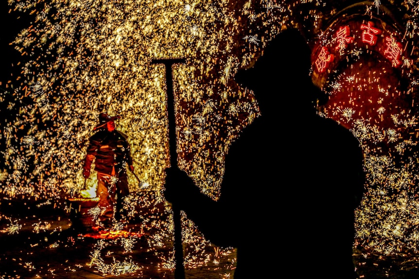 A man watches as his friend throws molten iron against a wall, creating sparks.