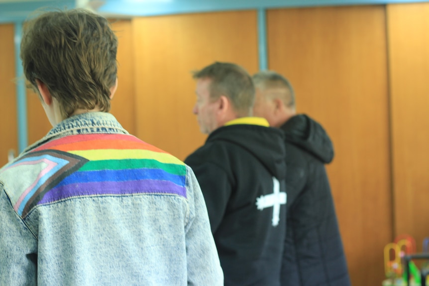 two people face away from the camera, one wears a rainbow pride flag on their denim jacket, the other wears a black union jumper