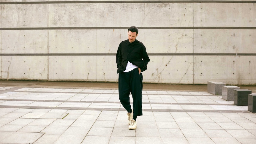 dance system (white man) walking through in a concrete open area, he's wearing black attire with a white tee undeneath