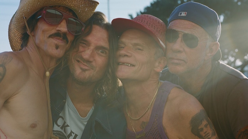Four members of Red Hot Chili Peppers stand in a line cheek-to-cheek and smiling. Three wear hats, two wear sunglasses.