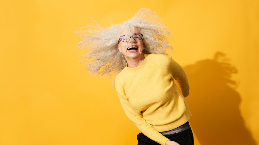 mature woman smiling and dancing wearing a yellow sweater against a yellow background