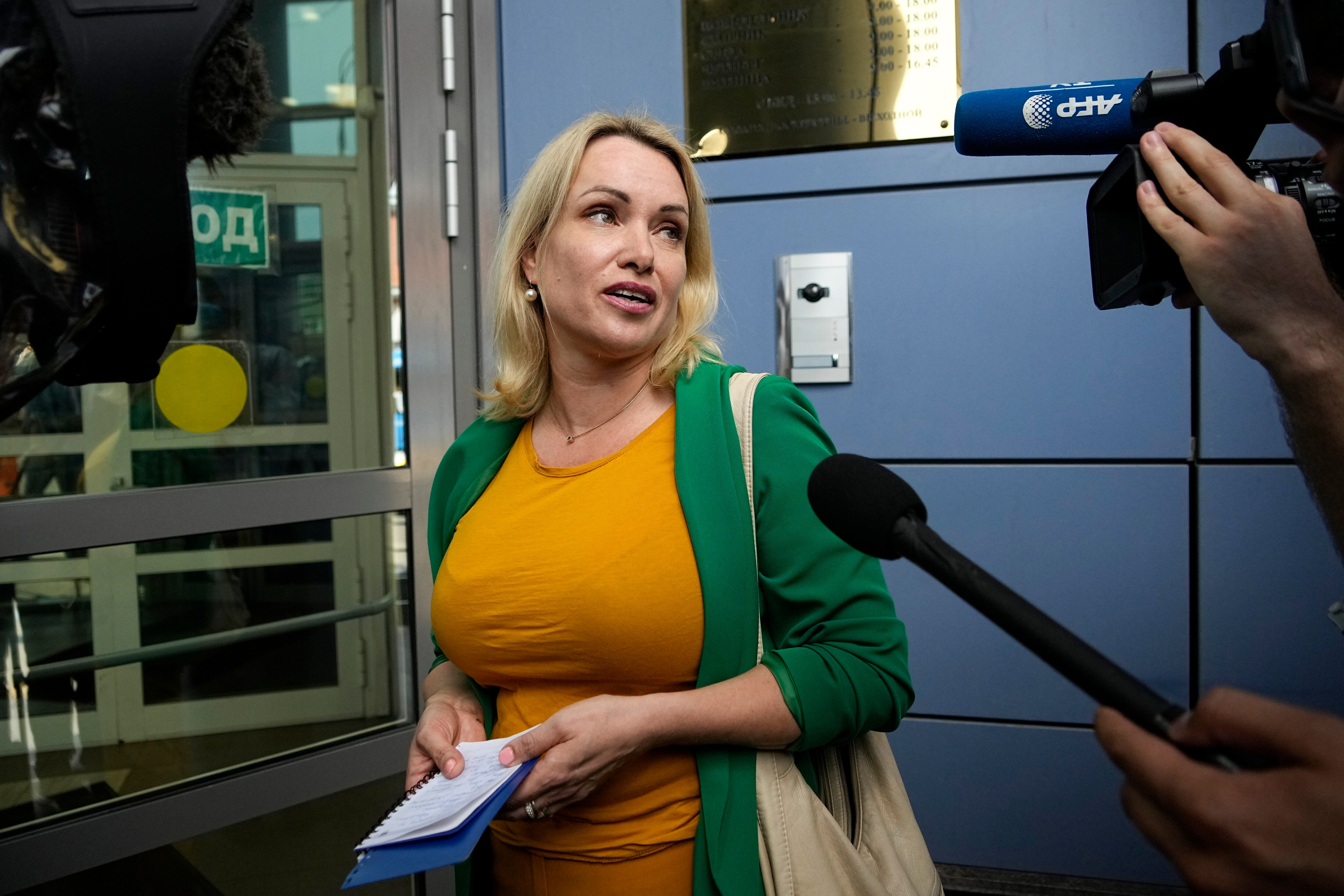 A blonde woman in an orange top and green jacket speaks to assembled media outside a court building door.