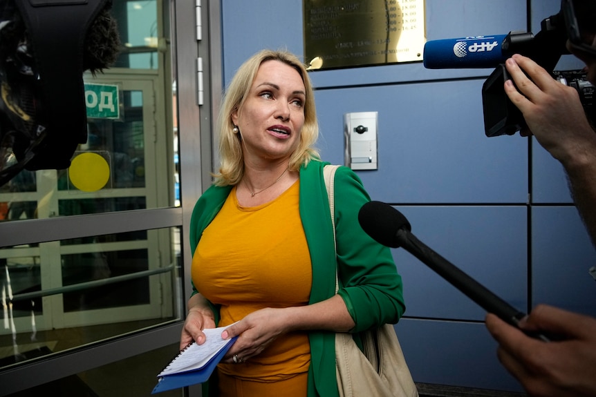 A blonde woman in an orange top and green jacket speaks to assembled media outside a court building door.