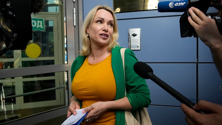 A blonde woman in an orange top and green jacket speaks to assembled media outside a court building door.