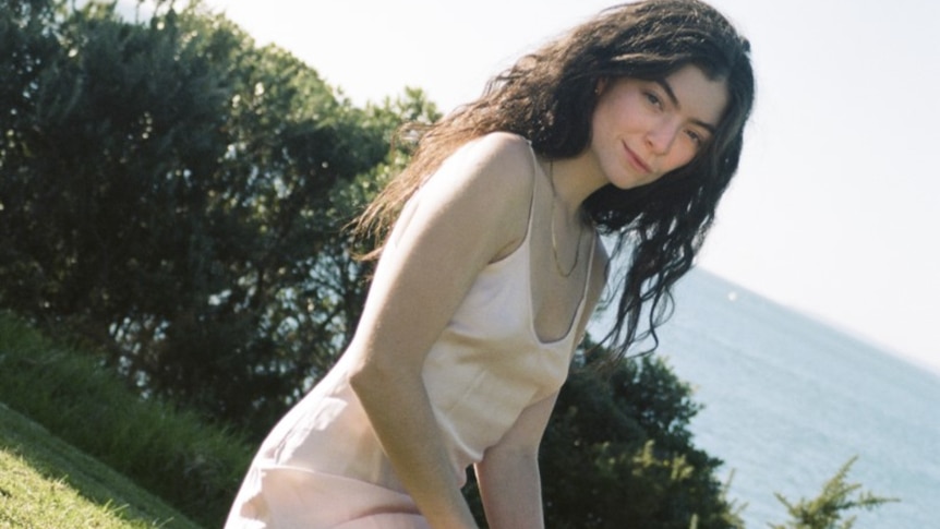 Brunette caucasian woman wears a peach coloured camisole dress, greenery and ocean behind her