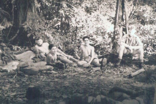 A black and white photo of four soldiers sitting on the ground and relaxing in the bush.