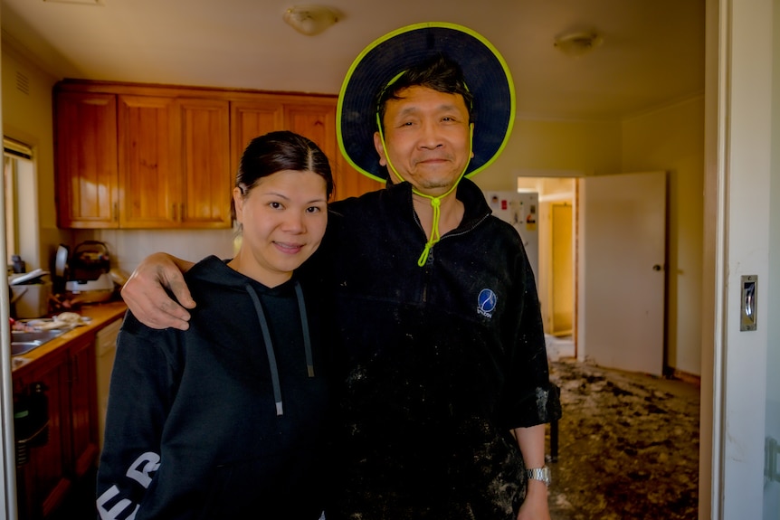 A couple smile and pose for a photo inside a flood-damaged kitchen