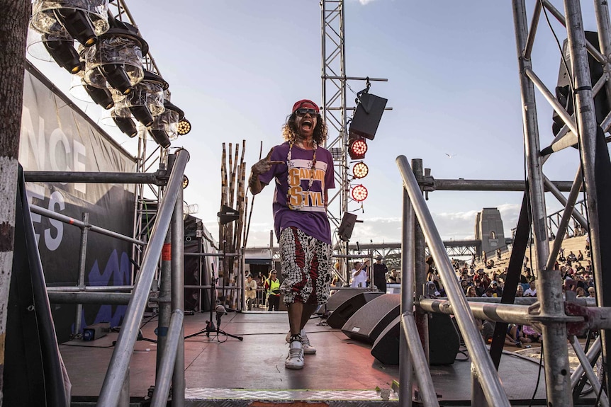 Colour photo of Sean Choolburra posing on the Dance Rites stage on the Sydney Opera House forecourt on a clear day.