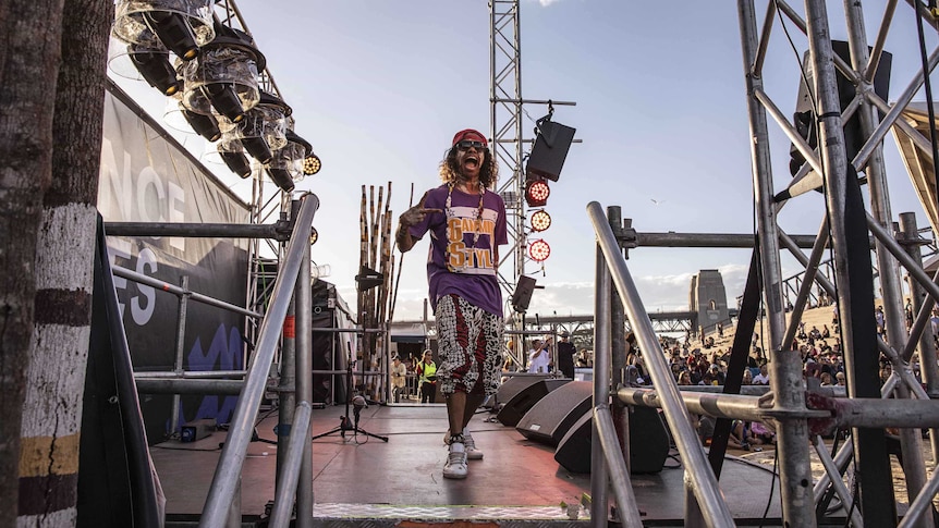 Colour photo of Sean Choolburra posing on the Dance Rites stage on the Sydney Opera House forecourt on a clear day.