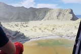 An emergency service worker sits in an open helicopter flying towards a bay where a deadly volcano eruption has just occurred