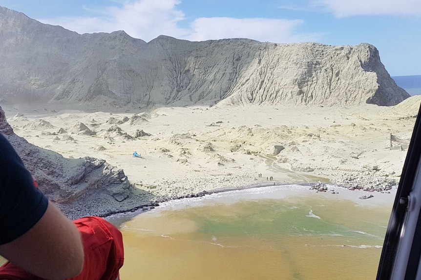 An emergency service worker sits in an open helicopter flying towards a bay where a deadly volcano eruption has just occurred