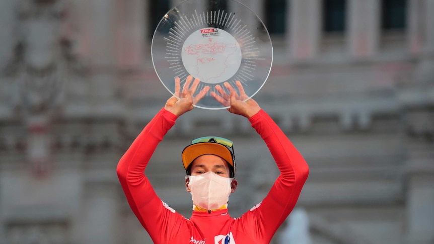 Primoz Roglic wears a red jersey and holds a round, see-through trophy in both hands above his head