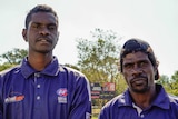 Two Aboriginal men in blue AFLNT shirts stand beside each other on an AFL pitch in the sun