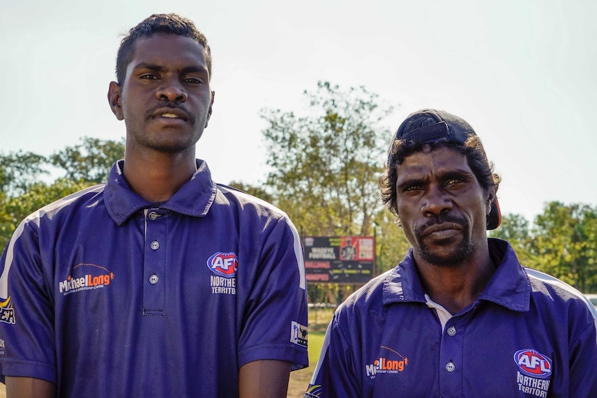 Two Aboriginal men in blue AFLNT shirts stand beside each other on an AFL pitch in the sun