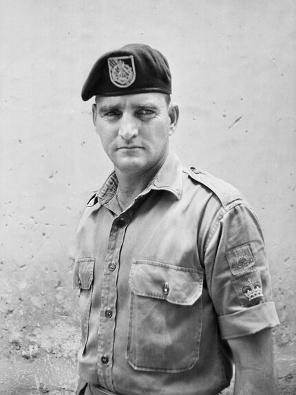 Soldier with short sleeves and beret stares unsmiling at camera with arms folded behind back