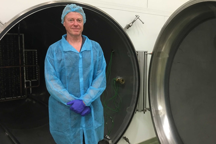 Michael Buckley stands in front of large circular dryer door dressed in hospital-style scrubs.