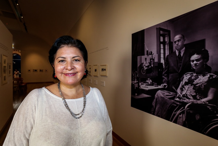 Museum director looks directly at camera. She stands beside a large portrait of Frida Kahlo and her doctor.