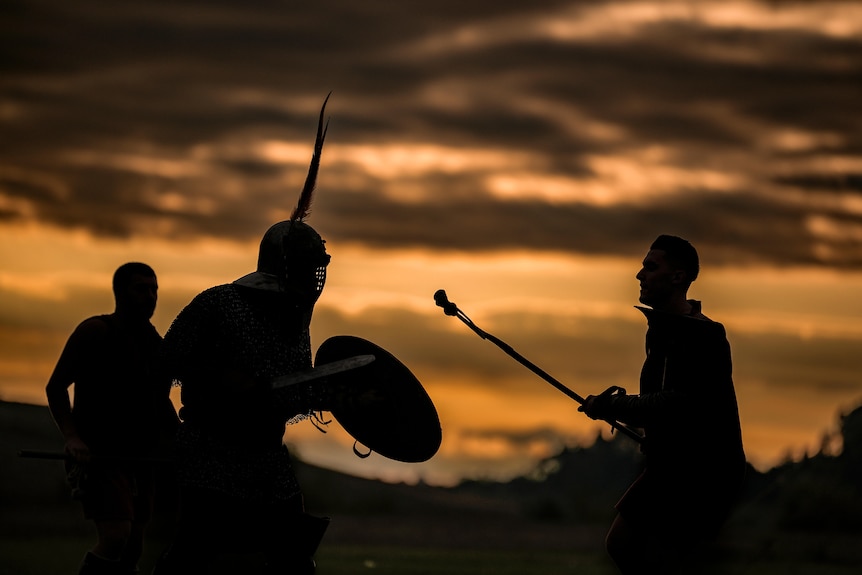 the silhouette of three men is shown against a cloudy yellow sky. Two of the men re-enact a gladiator battle
