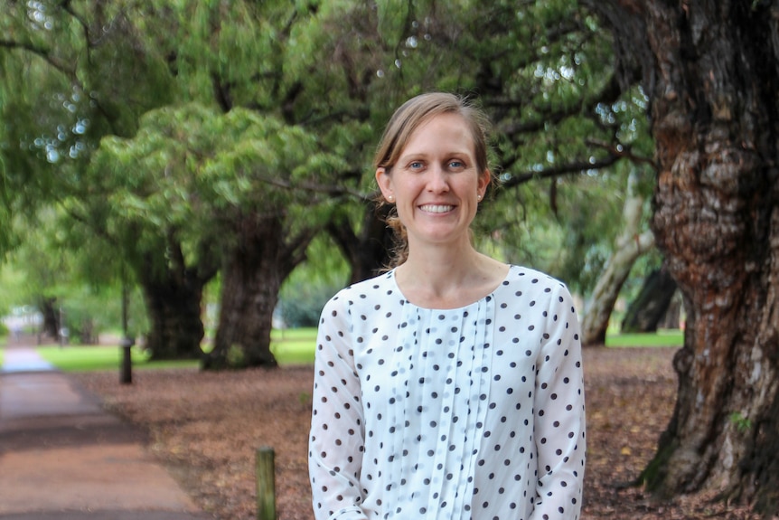 A middle-aged woman in a white patterned top smiles widely, stands in front o trees and a path.Brown hair tied back.