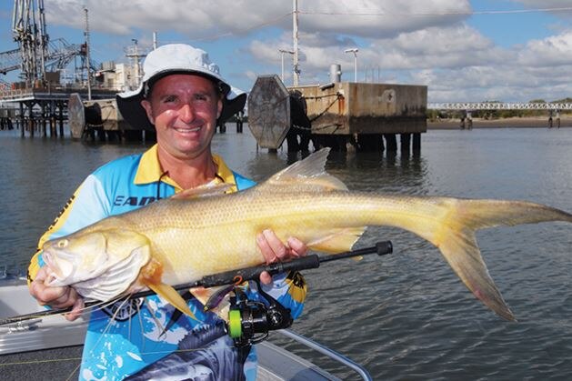 A man in a blue and yellow fishing shirt holding a large yellow and silver fish next to his fishing rod.