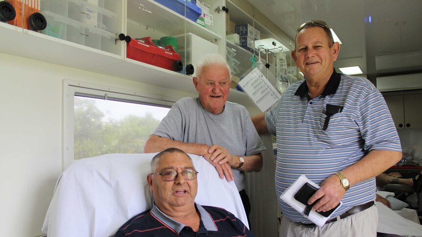 Three men stand in a dialysis unit, one is dialysing in a chair and the other two are standing next to him.