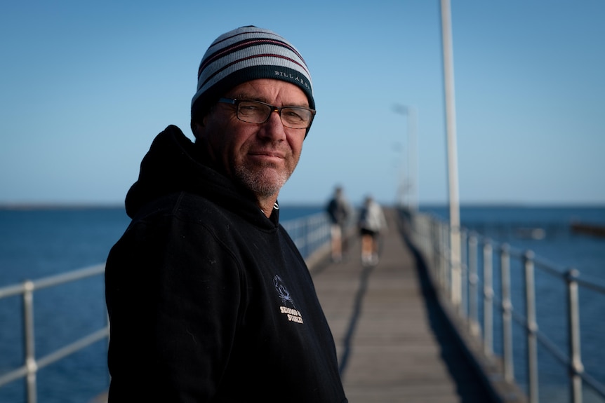 A man wearing a jumper and beanie stands on a jetty side on and looks at the camera with a serious expression