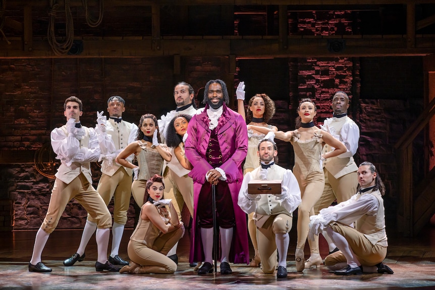 Young man in 18th century coat, bright pink satin, standing on stage smiling, with other performers posing around him.