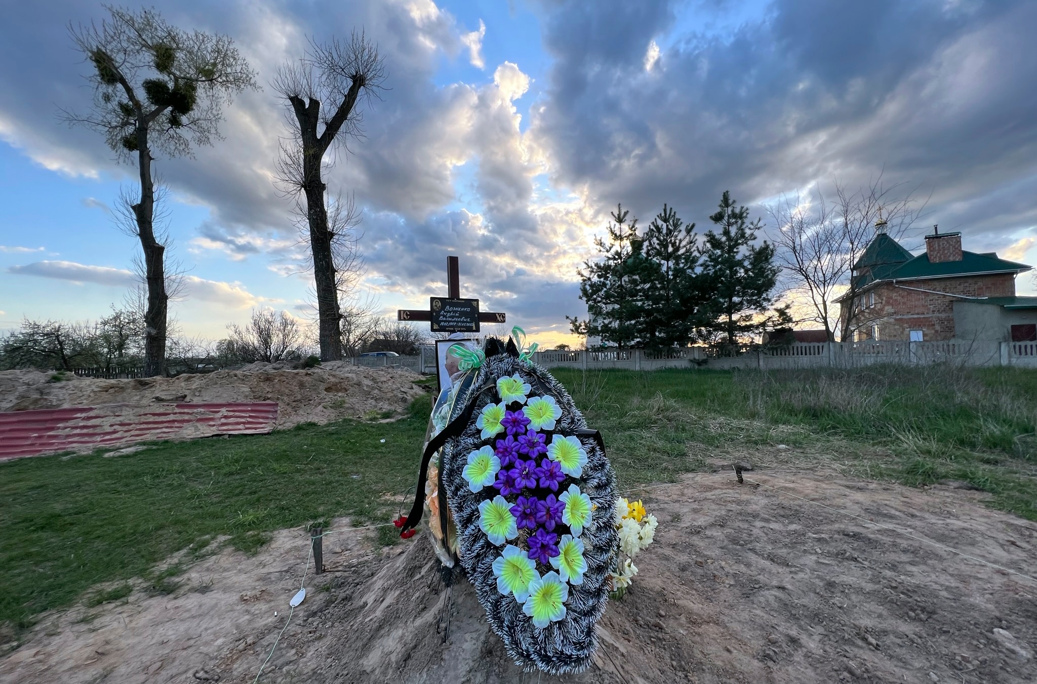 A floral garland sits on top of a grave in a field near a brick home. 