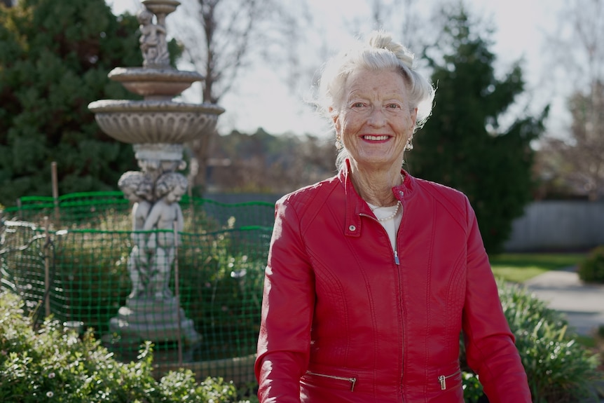 Elderly woman wearing a red jacket, standing outside in a garden.