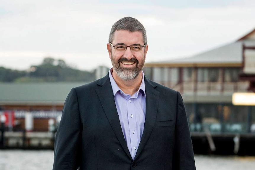 Man in suit stands in front of Perth rowing club and river