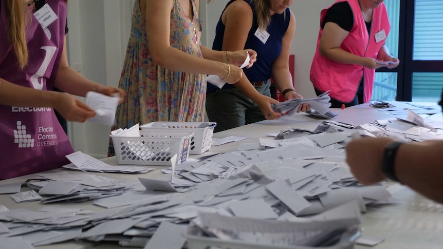 Four Electoral Commission Queensland staff count ballot papers, ballot papers scattered all over table.