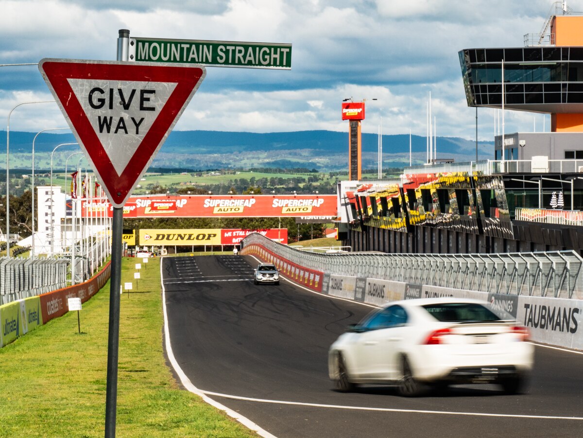 Give way Mountain Straight street signage in the foreground with cars driving along a race circuit.