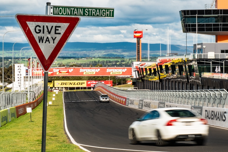 Give way Mountain Straight street signage in the foreground with cars driving along a race circuit.