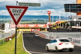 Give way Mountain Straight street signage in the foreground with cars driving along a race circuit.