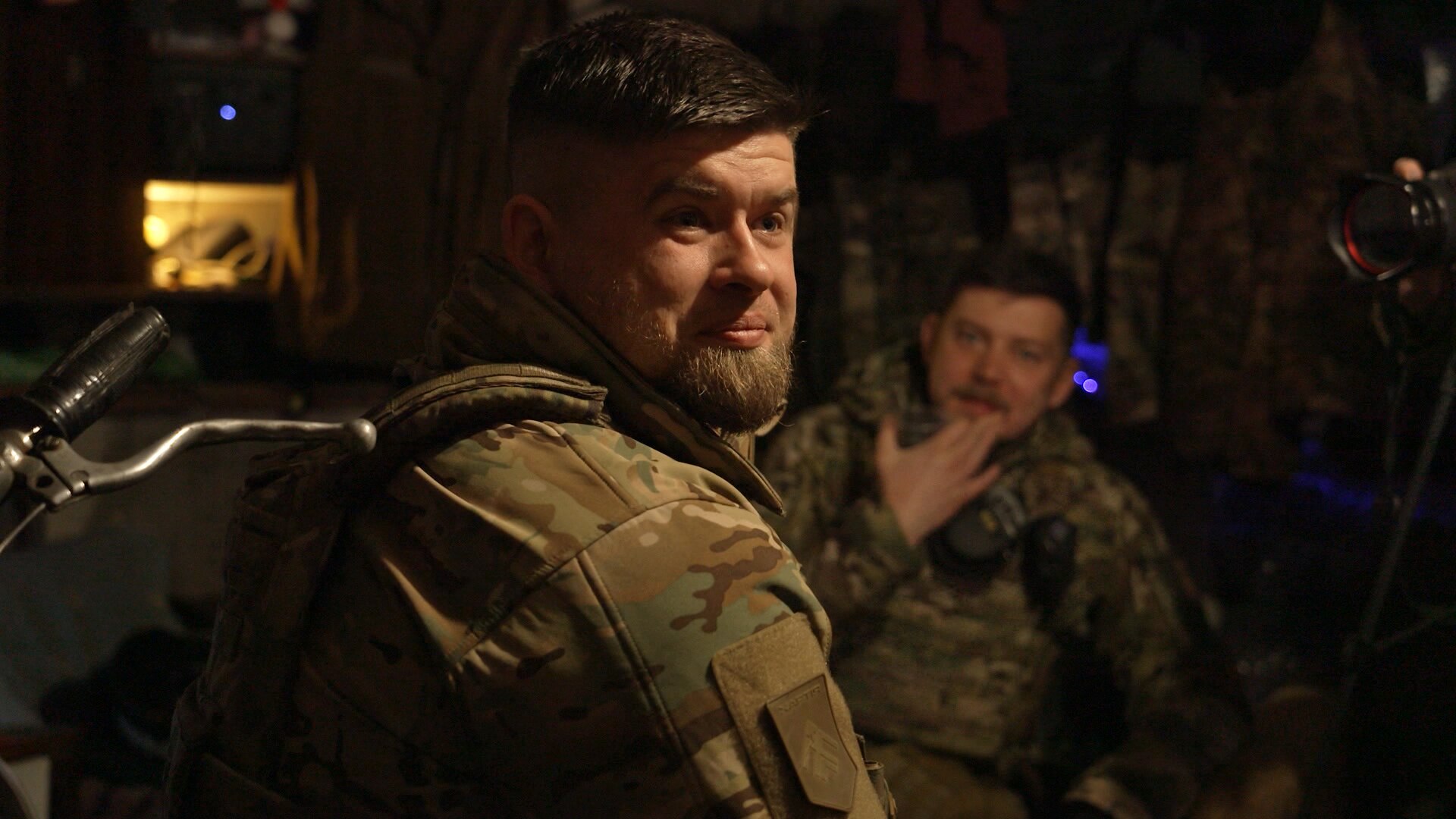 A man wearing a military uniform sits in a darkened room, smiling. Another soldier sits nearby.