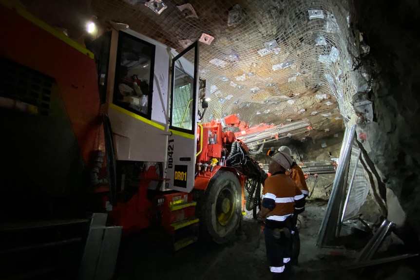 Two workers stand in an underground tunnel next to a large drill, surveying the mesh and bolts on the walls.