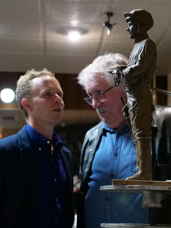 Two men stand talking behind a small model of a statue of a jockey.
