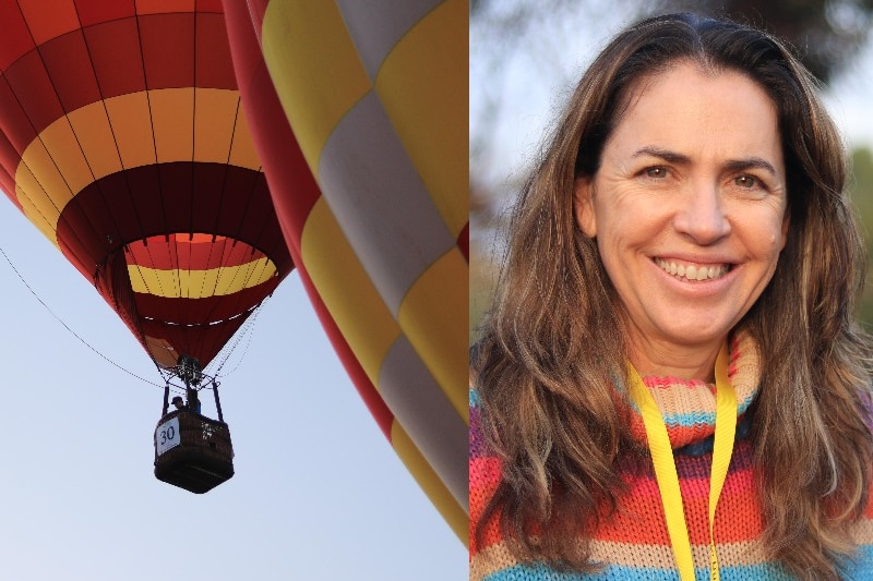 A woman smiles and wears a colourful jumper next to hot air balloons in the sky.