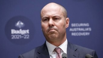 Josh Frydenberg looks to the left as he stands in front of a blue budget background. He wears a navy suit and pink tie.