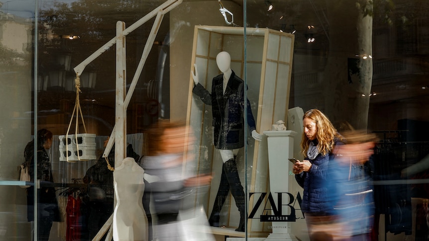 People walk by a Zara shop window, with a white mannequin in a packing crate wearing a black jacket and boots