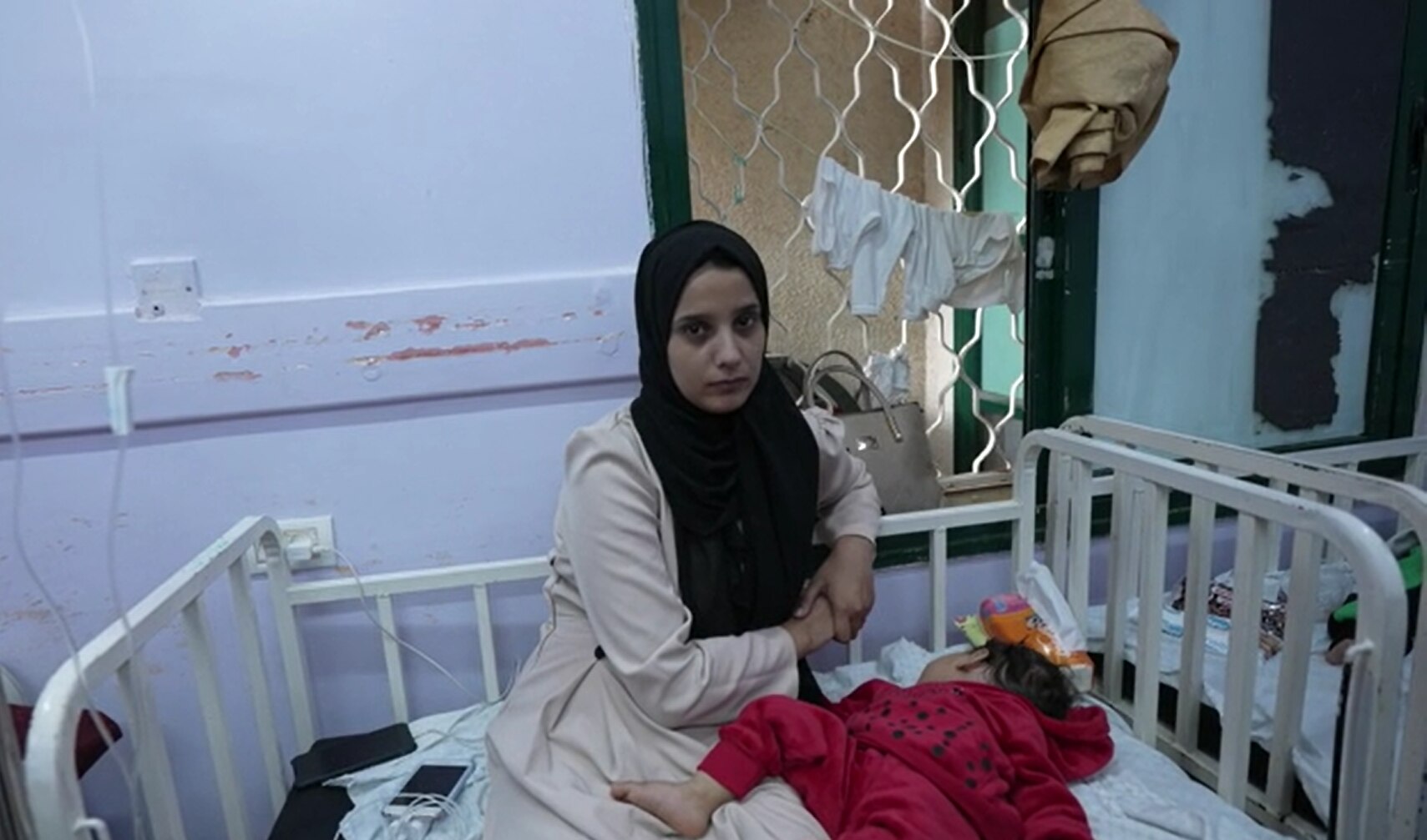 A woman wearing a black hijab is seen with her baby sitting on a hospital bed. 