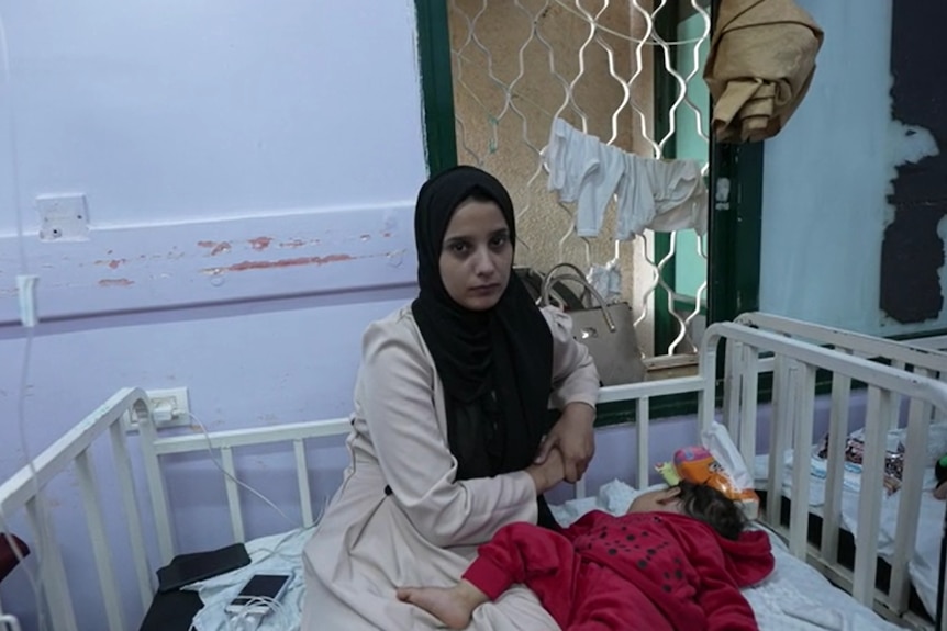 A woman wearing a black hijab is seen with her baby sitting on a hospital bed. 