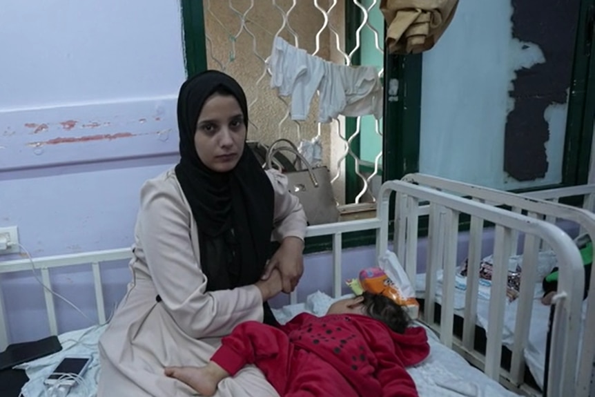 A woman wearing a black hijab is seen with her baby sitting on a hospital bed. 