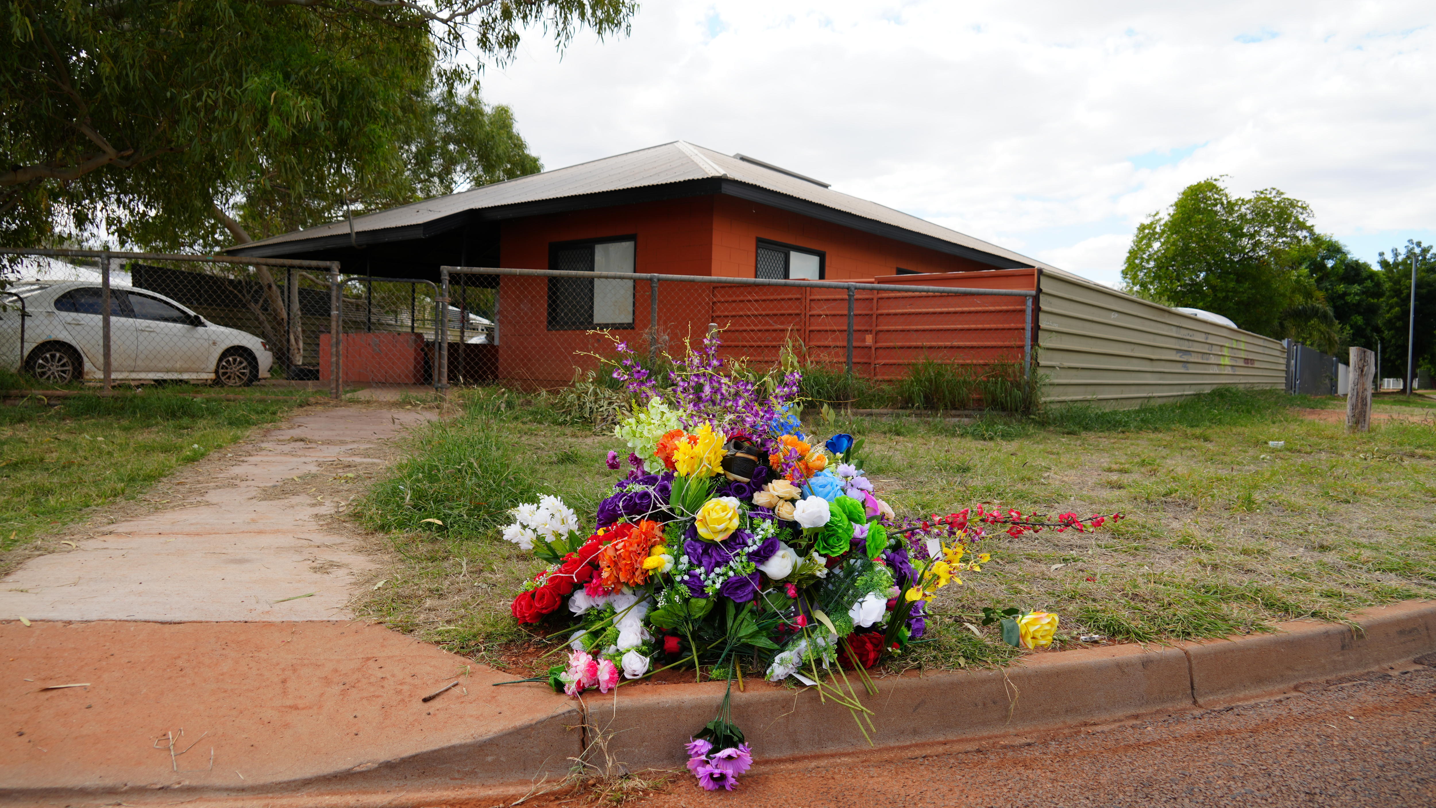 A wide-shot of multi-colour flower memorial resting on the edge of a curb in front of a home, with orange paint.