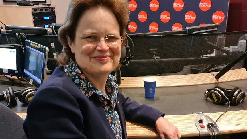 woman with glasses, blue jacket and floral blouse smiles, elbow resting on studio desk at ABC