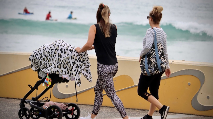 Two woman walk along a path in front of the beach, one of them is pushing a pram.
