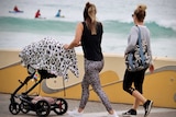 Two woman walk along a path in front of the beach, one of them is pushing a pram.