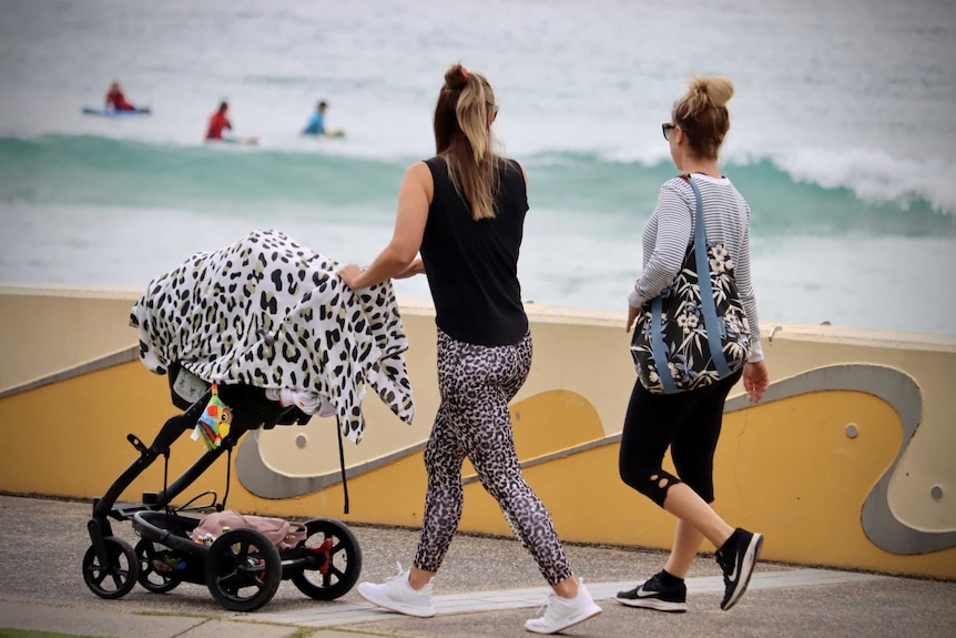 Two woman walk along a path in front of the beach, one of them is pushing a pram.