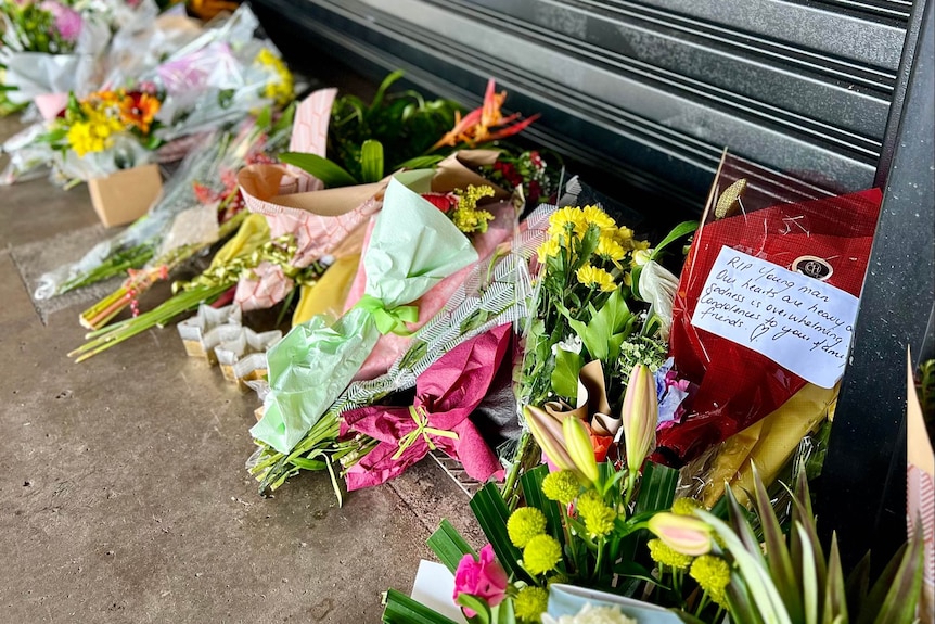 A row of bunches of flowers lying on the pavement, in front of a metal roller-door. 